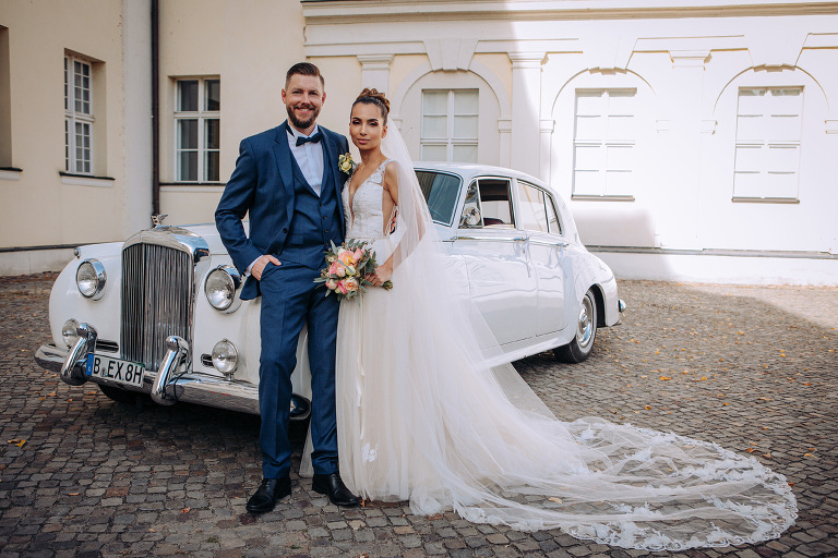 Die Braut und Bräutigam posieren vor dem Hochzeitsauto im Schloss Köpenick